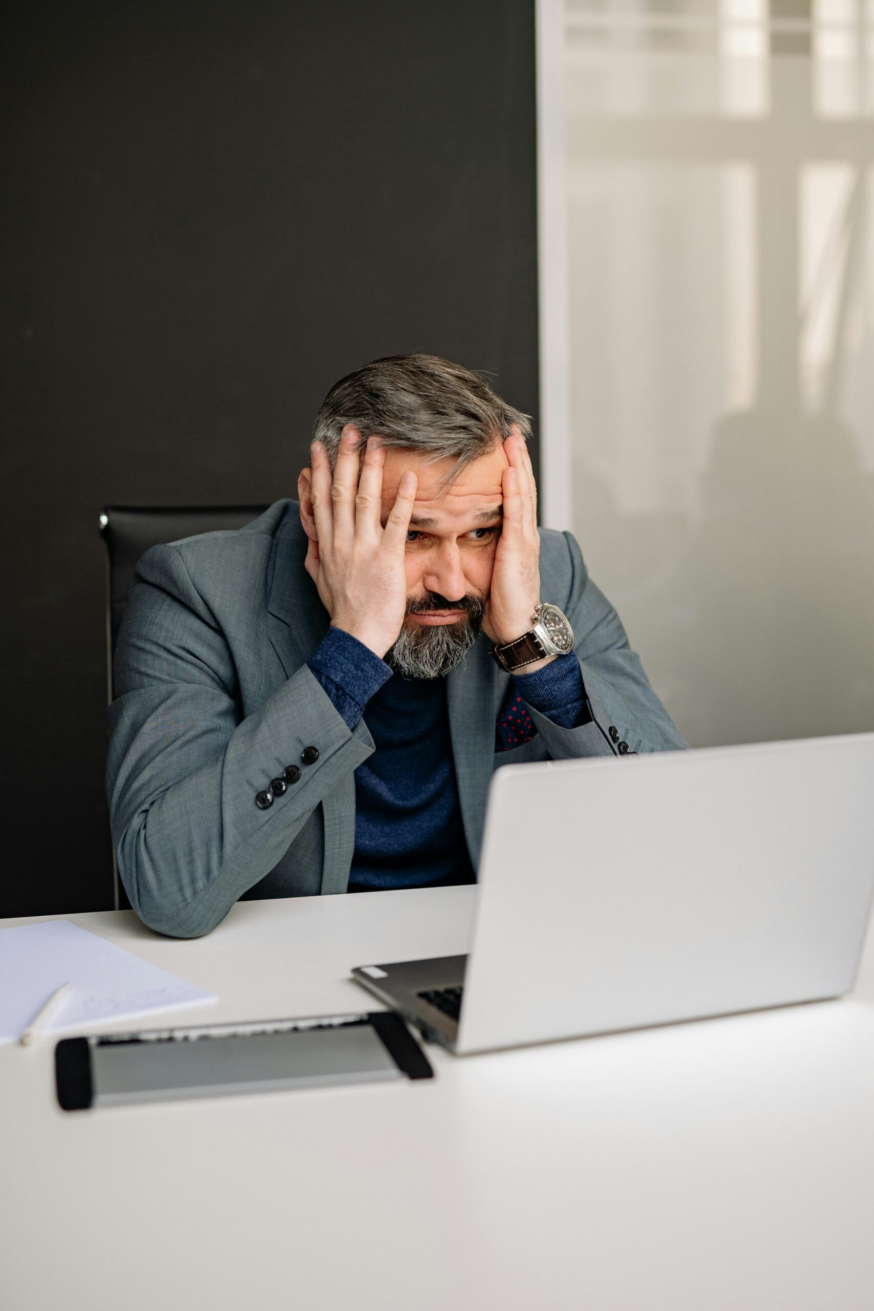 A stressed adult man in a suit at an office desk with hands on face and laptop.